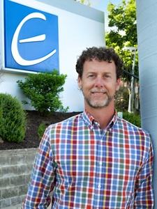 Half-body photo of Monte Cooper, a Caucasian man with short curly brown hair and a brown and white beard. The JEA logo of a white letter E on a blue background is on the wall behind him.